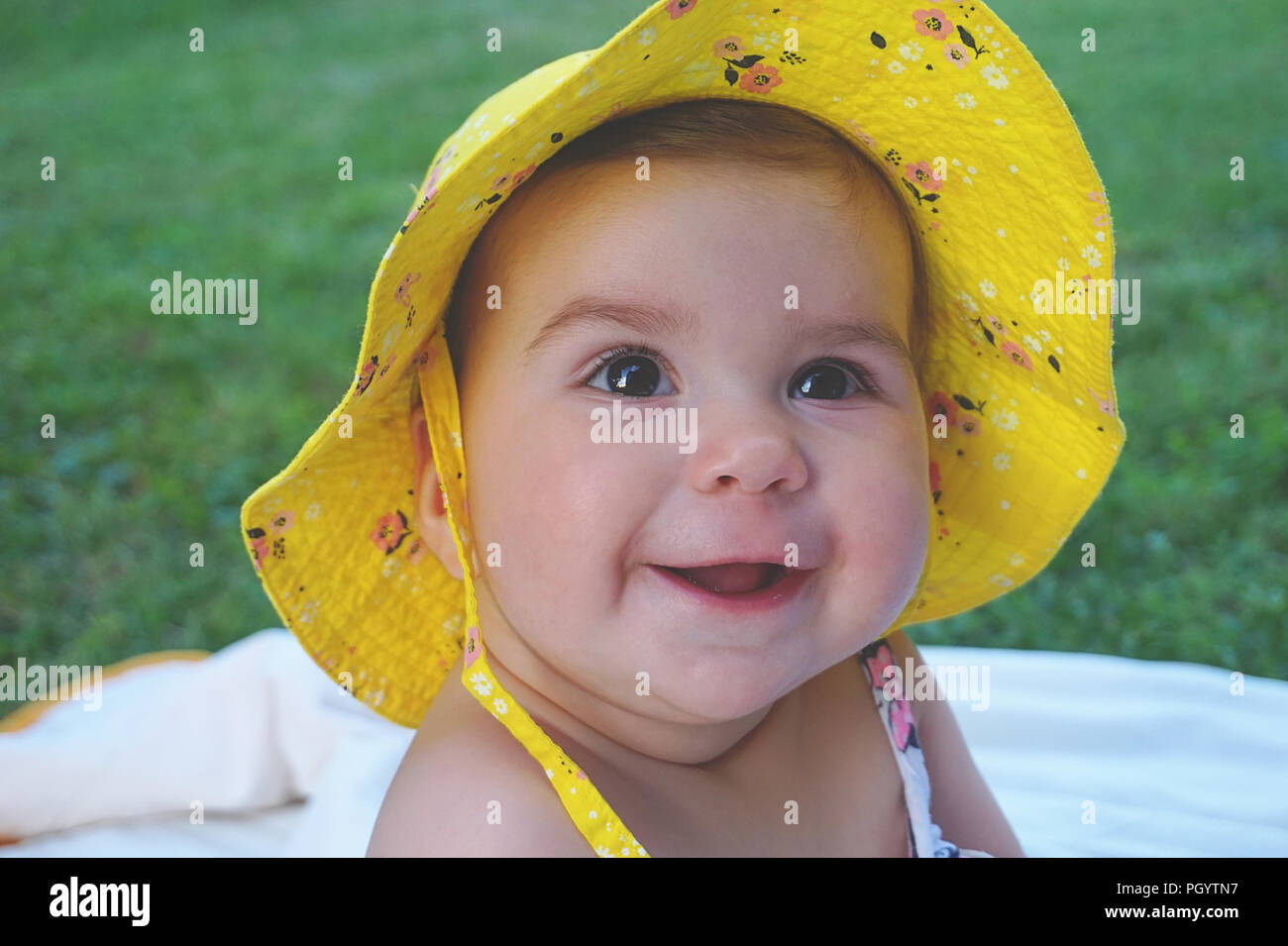 Portrait of cute and happy beautiful baby girl with yellow hat smiling and looking up on the green meadow Stock Photo