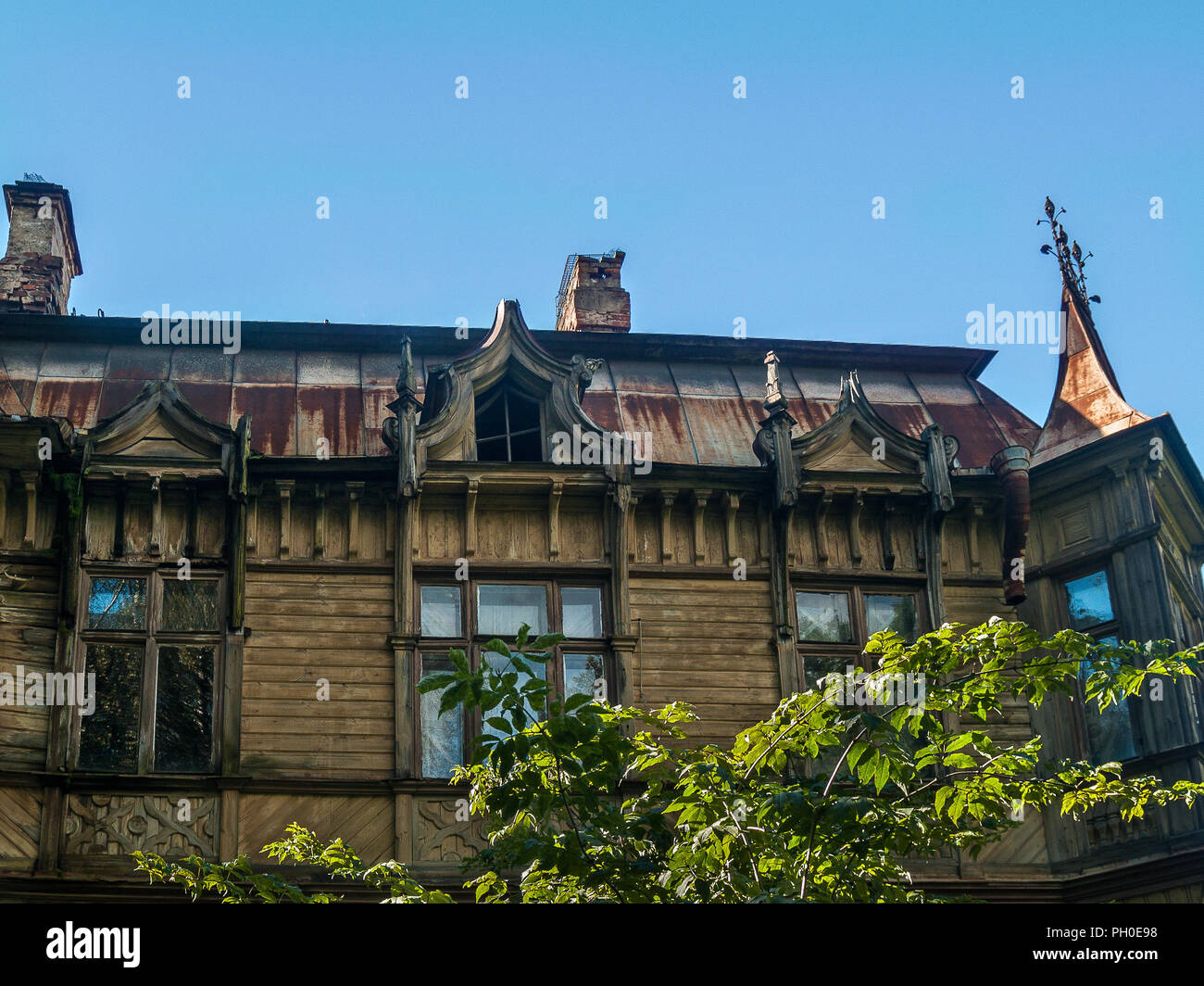 The facade of the wooden house in eclectic style with a bay window, carved attic windows, figured platbands, with a galvanized rusty roof on a clear s Stock Photo