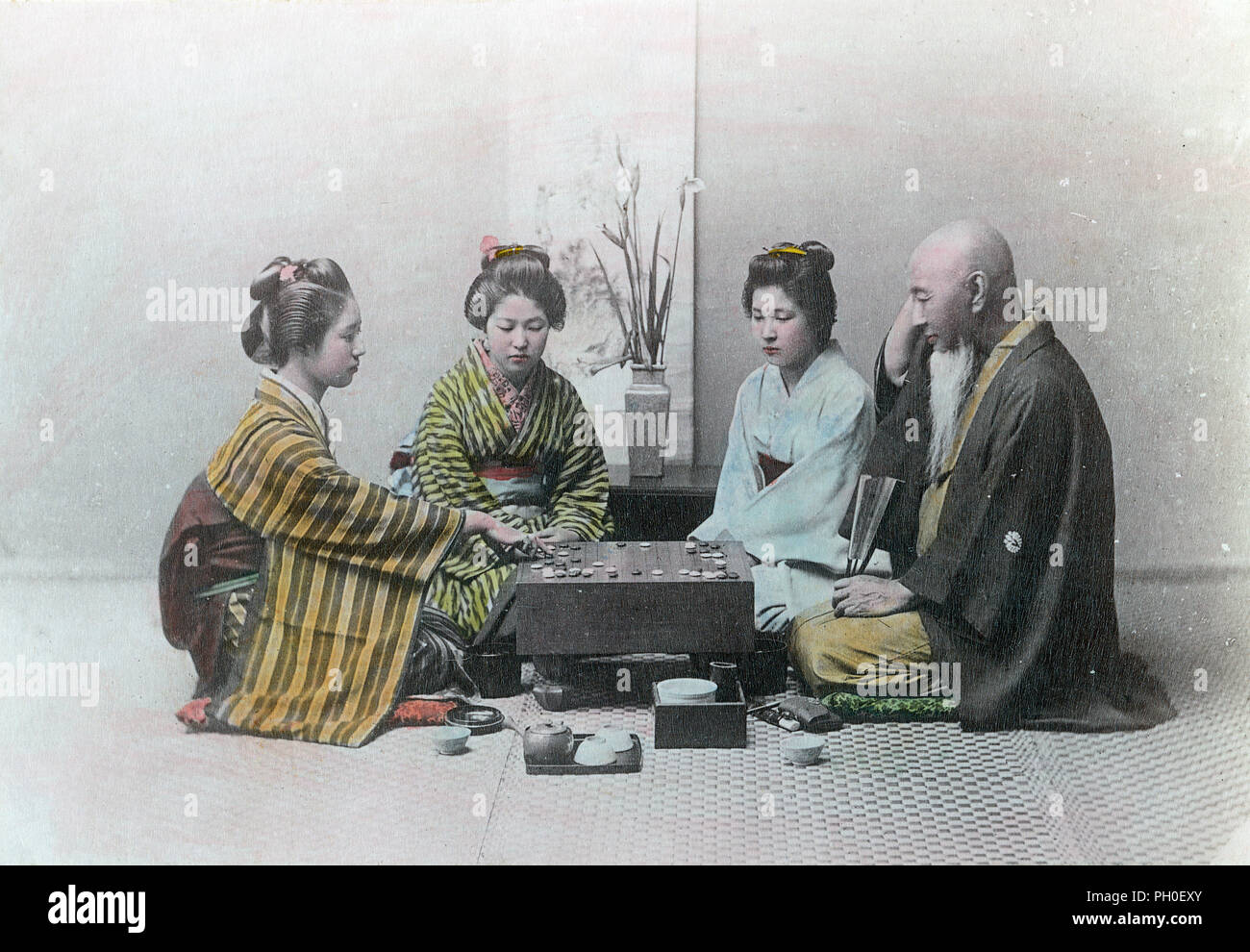 [ 1890s Japan - Playing Go ] —   Three young women in kimono and traditional hairstyles and an elderly man in a haori coat are seated around a board for the game of go.  This studio is arranged as a zashiki (guest room), with an ikebana flower arrangement and scroll in the back. A small tray with a teapot and two cups can be seen at the front.  19th century vintage albumen photograph. Stock Photo