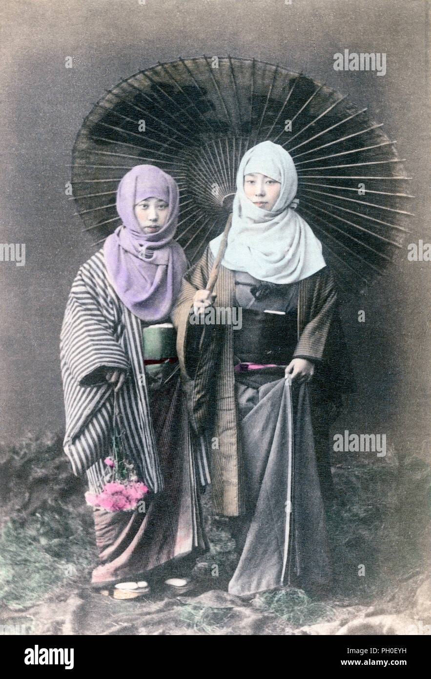 [ 1890s Japan - Women in Winter Clothes ] —   In this studio photo, two women in thick winter kimono have okosozukin wrapped around their head to protect themselves against the cold. One of the woman is wearing a parasol, the other some flowers.  19th century vintage albumen photograph. Stock Photo