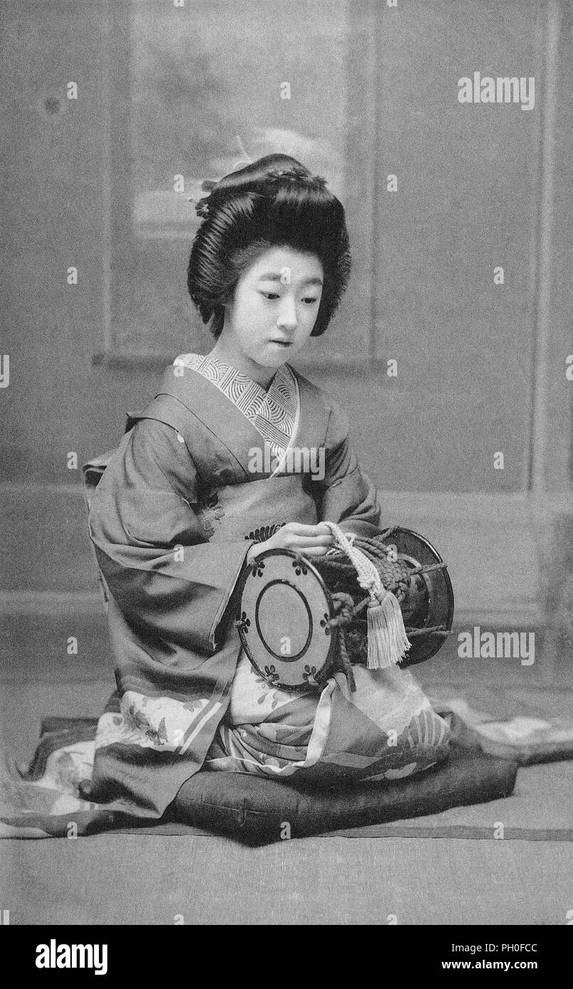 [ 1920s Japan - Geisha with a Tsutsumi Drum ] —   A geisha in kimono and traditional hairstyle is holding a Tsutsumi (also Tsuzumi) shoulder drum. The same type of drums were used in Noh and Kabuki dramas.  The Tsutsumi is held with the left hand to the right shoulder, while the right hand is used to strike. The instrument has two drumheads supported by tension cords. These cords are pulled to change the pitch.  20th century vintage postcard. Stock Photo