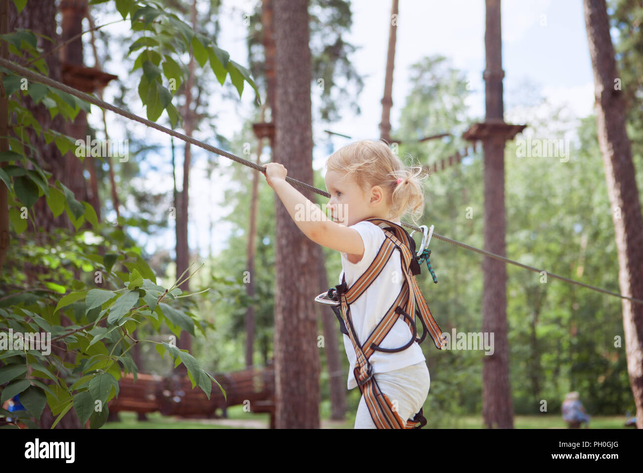 Child girl in safety harness attached with carbine to cable moves carefully along rope way. Stock Photo