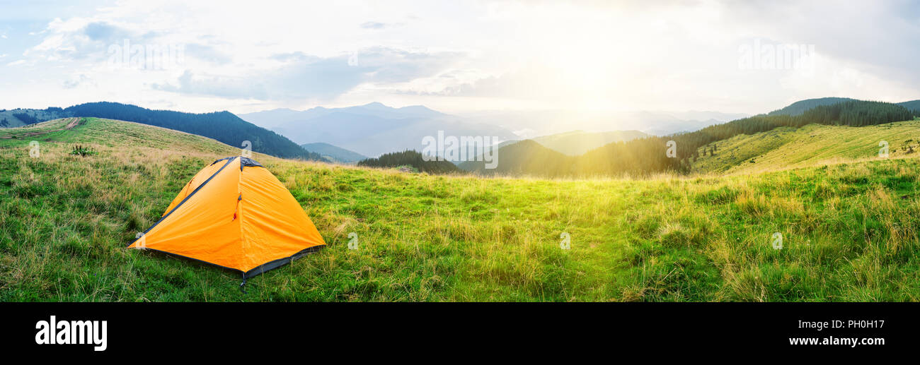 Orange tent on meadow with green grass in mountains under bright Stock Photo