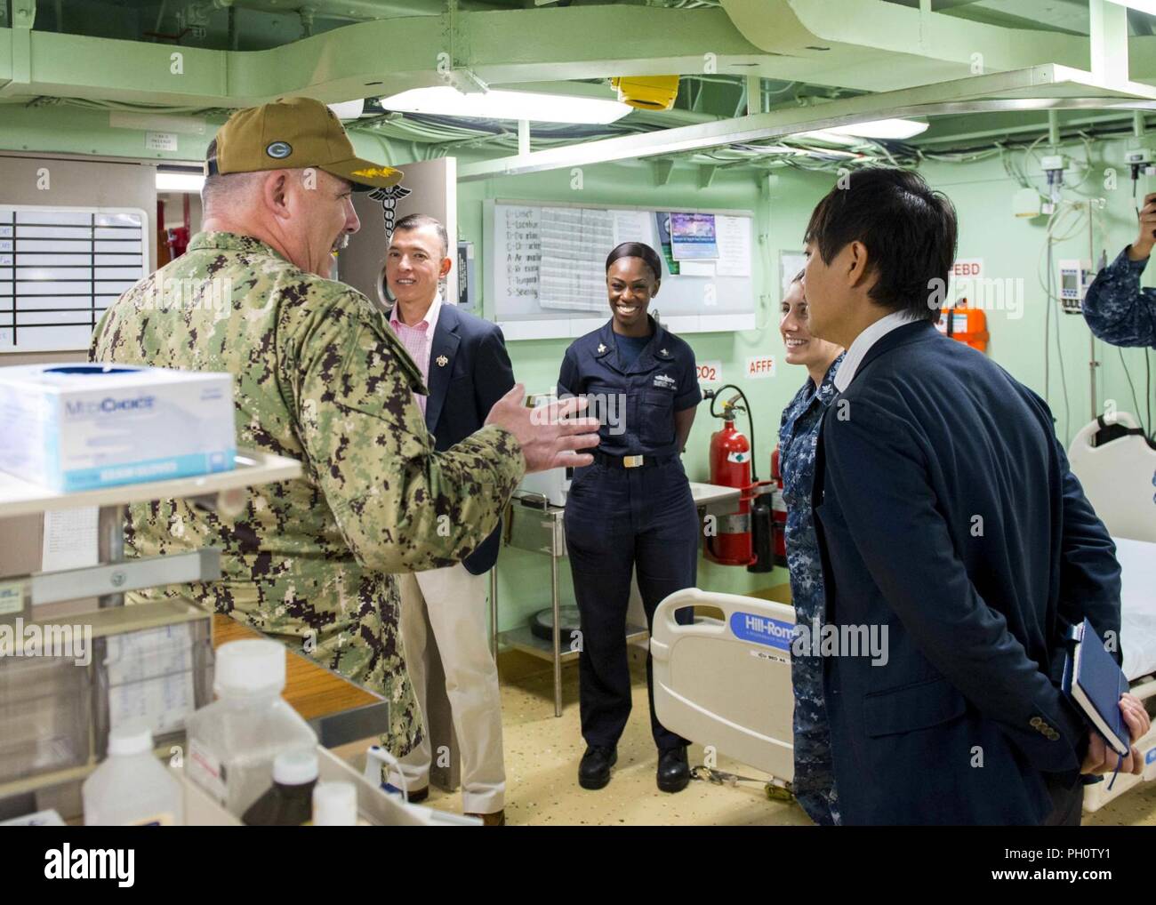 Japan (June 21, 2018) Capt. Michael Harris, executive officer of the San Antonio-class amphibious transport dock ship USS Green Bay (LPD 20), gives a tour of the medical bay to Takahiro Suzuki, Ministry of Foreign Affairs Status of Forces Agreement deputy director, North American Affairs Bureau, aboard Green Bay, June 21, 2018. The tour of CFAS and the ship was given to increase his understanding of CFAS’ capabilities in support of the U.S.-Japan alliance. Stock Photo