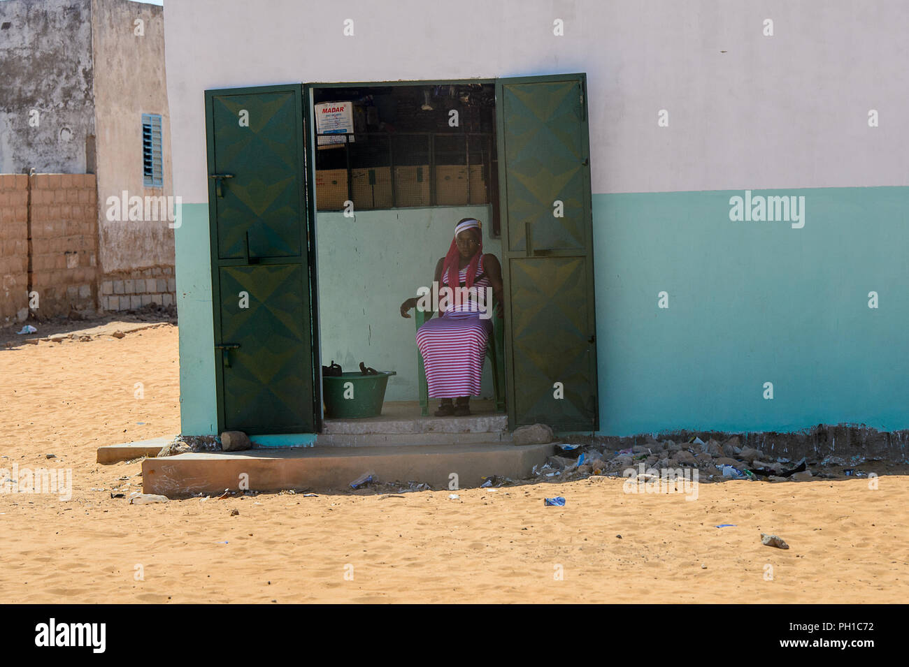ROAD TO LAMPOUL, SENEGAL - APR 23, 2017: Unidentified Senegalese woman in striped dress sits in a chair in the doorway. Still many people in Senegal l Stock Photo