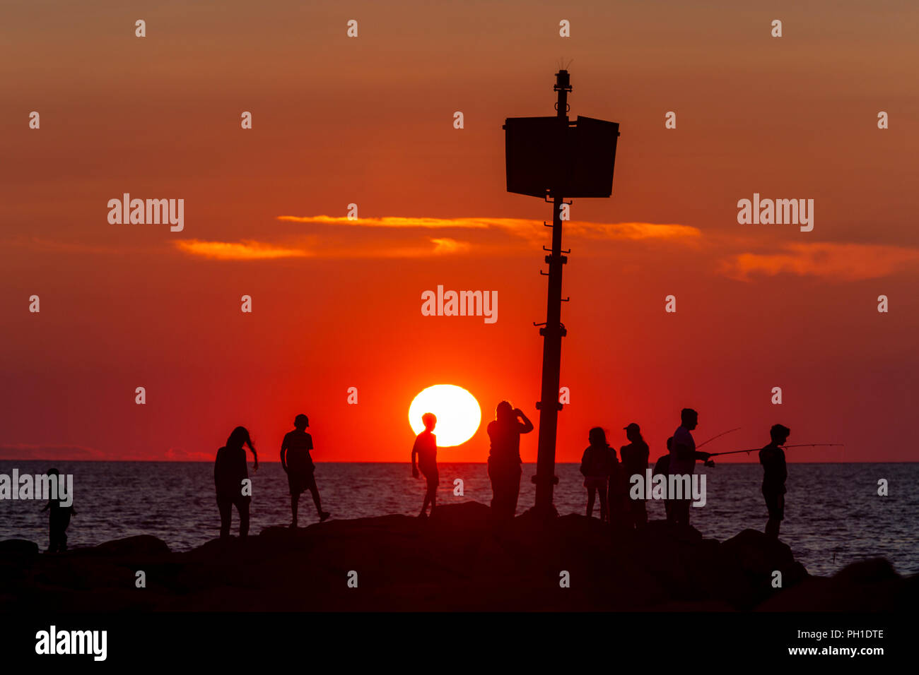 People stand on the jetty, some fishing, and watch the sunset at
