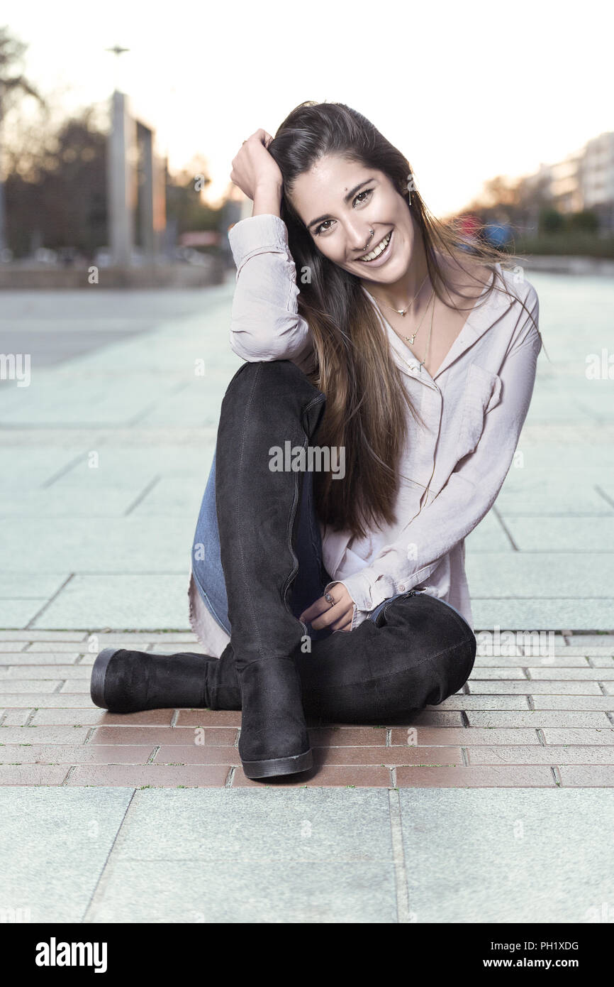 Beautiful girl in boots sitting on the street floor, with her head resting on her hand and smiling at you. Stock Photo