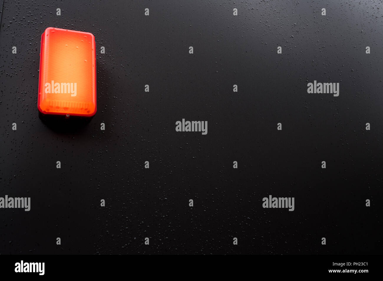 orange lamp suspended on a black wall. Image taken following rain so there are water droplets on the black wall Stock Photo