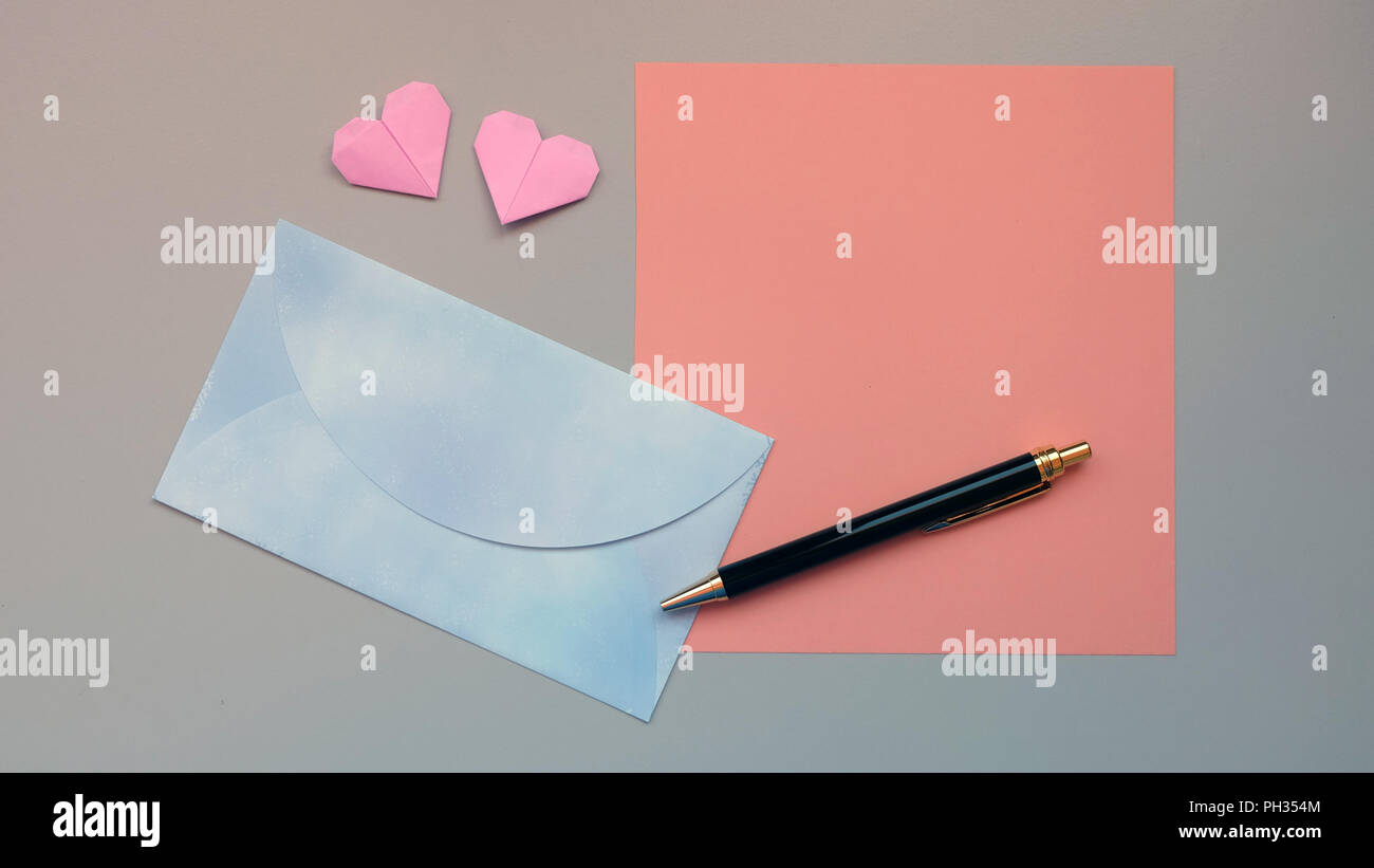 Flat lay of a blue envelope, a blank pink paper, black ball pen and a pair of pink folded hearts Stock Photo