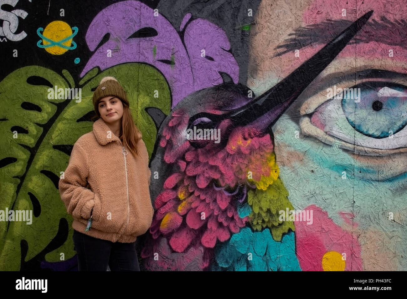 Young girl wearing a pom pom hat in front of a colourful bird art graffiti Stock Photo