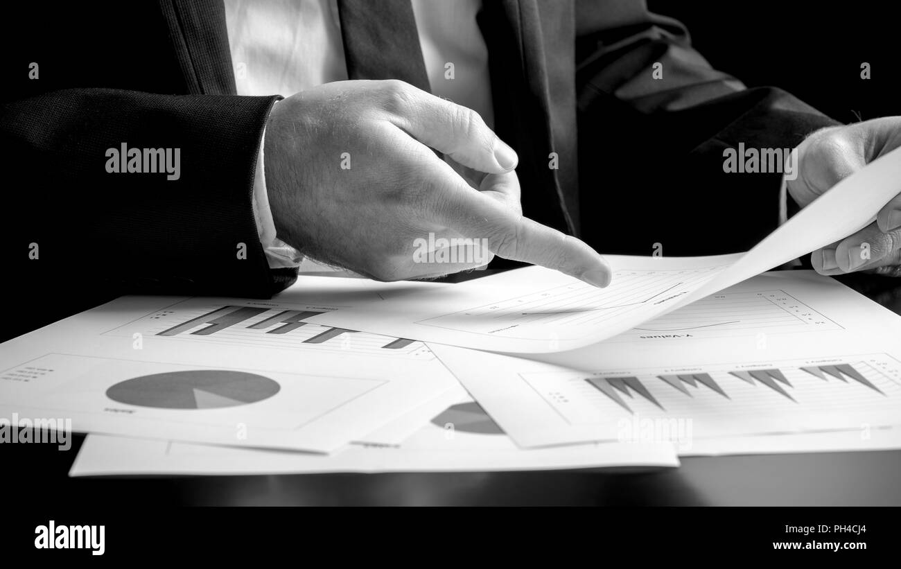 Businessman analysing a set of bar, pie and triangular graphs as he sits at his desk pointing to one document he is holding, close up greyscale in a b Stock Photo