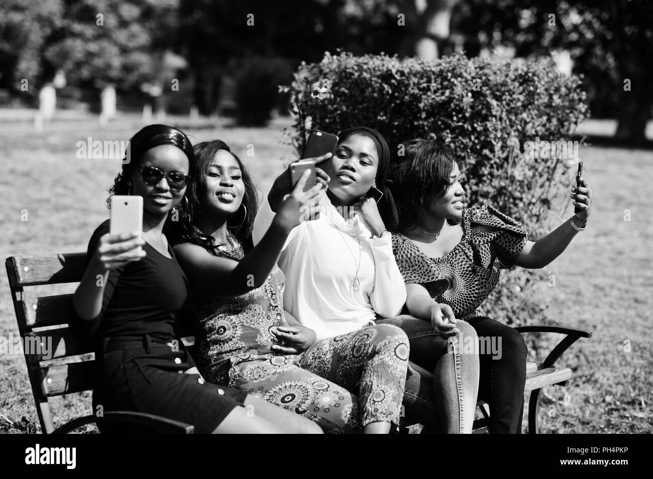 Group of four african american girls sitting on bench outdoor with mobile phones at hands and making selfie. Stock Photo