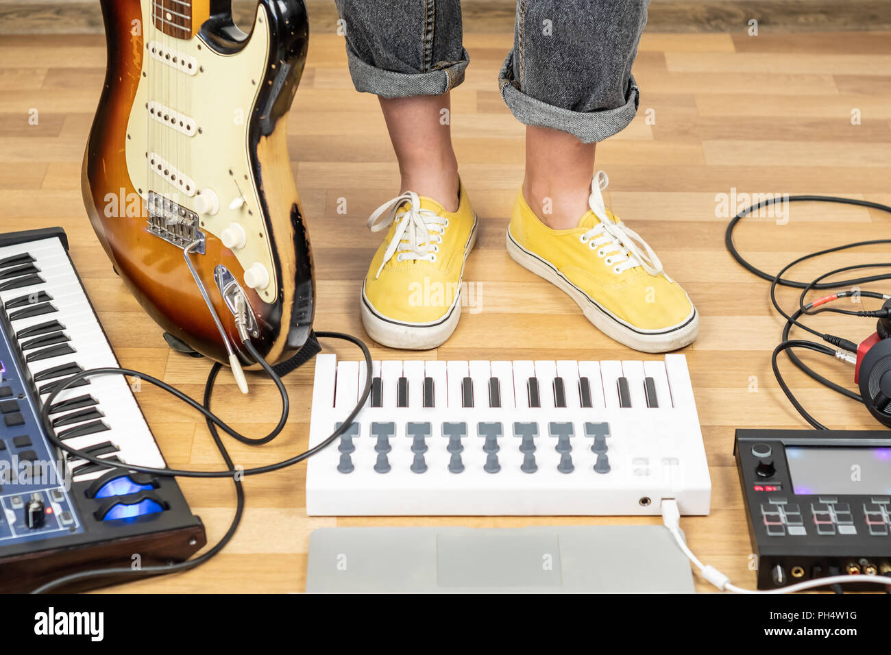 Indie musician with guitar and synthesizers at a rehearsal room. Young female guitarist in yellow shoes in home studio, close-up view of legs in yello Stock Photo