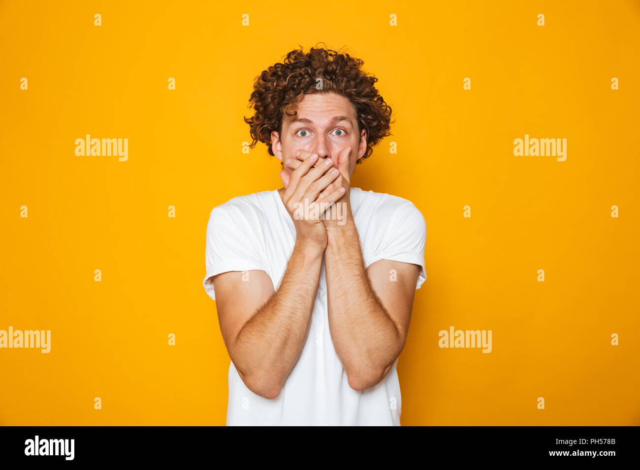 Portrait of a shocked curly haired man covering mouth isolated over yellow background Stock Photo
