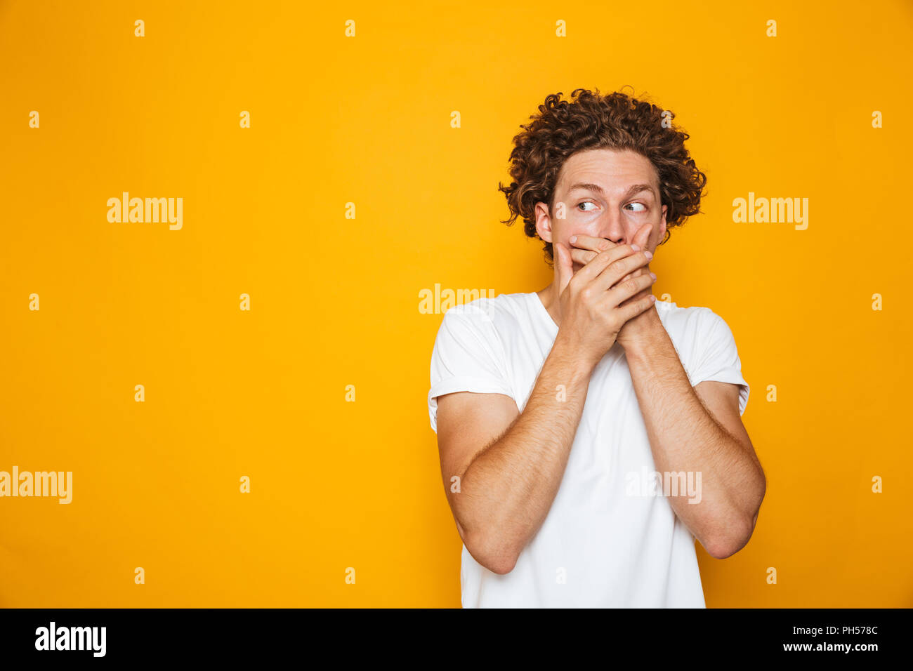 Portrait of a shocked curly haired man covering mouth isolated over yellow background, looking away Stock Photo