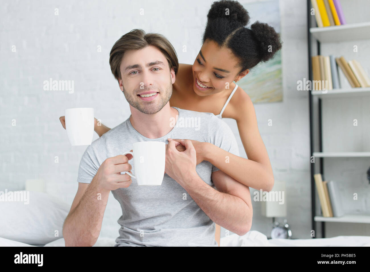 portrait of smiling multiracial couple with cups of coffee in bed in ...