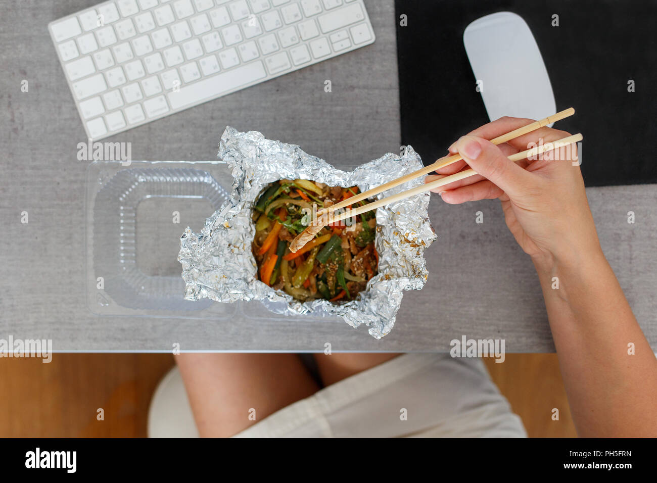 Young woman eating warm salad ordered in food delivery company. Chinese cuisine at the working desk Stock Photo