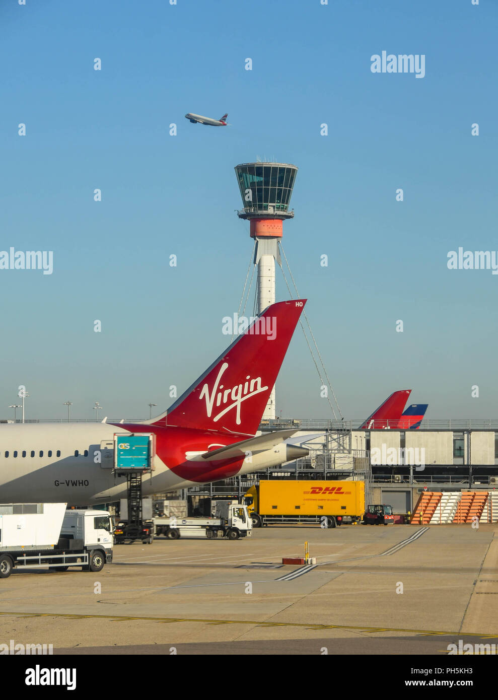 Tail fin of a Virgin Atlantic jet at London Heathrow airport. The airport's control tower and a jet taking off are in the background Stock Photo