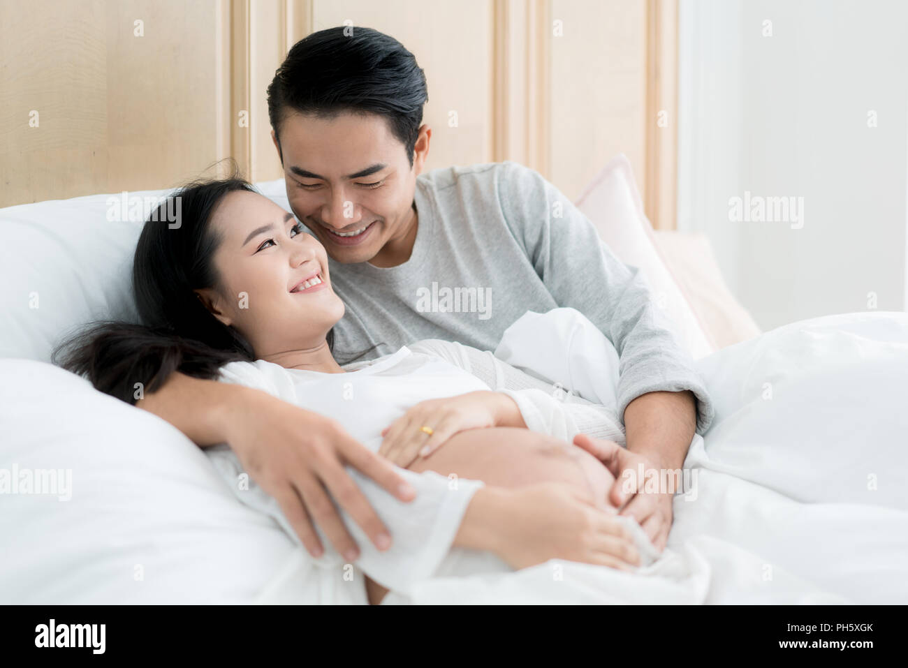 Handsome Asian man and his beautiful pregnant wife are hugging and smiling while lying in bed at home. Stock Photo