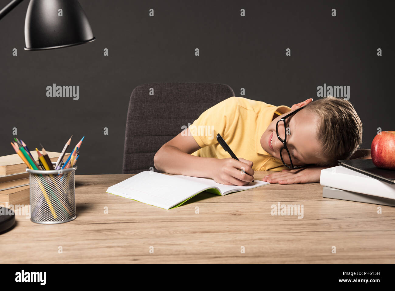 schoolboy in eyeglass laying on table and doing homework at table with lamp, books, colour pencils and textbook on grey background Stock Photo