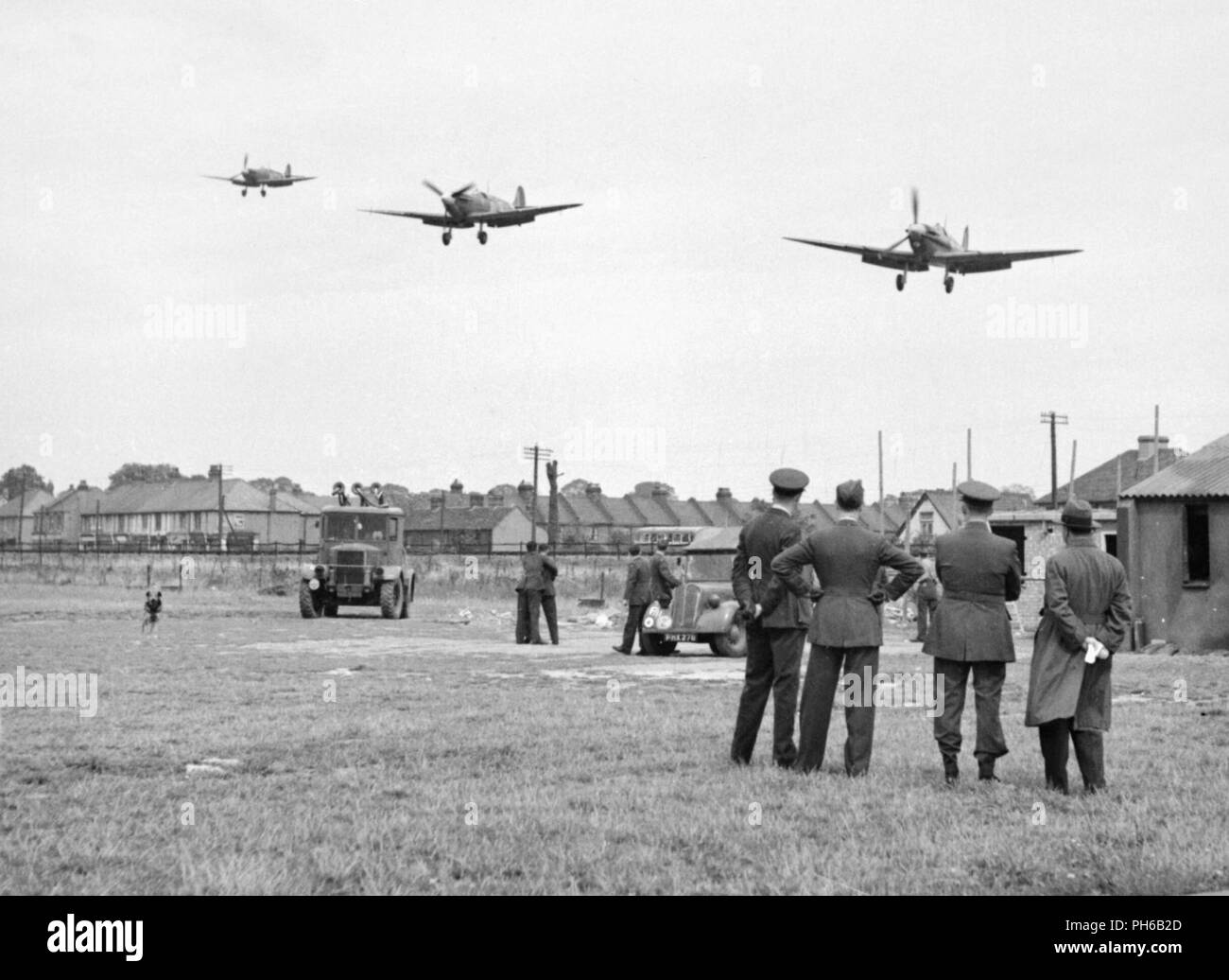 Personnel of No.121 (Eagle) Squadron look on as three Supermarine Spitfire aircraft land after a fighter sweep over northern France at Royal Air Force Rochford in Essex, England August 1942. Some of the accommodation used by the squadron is visible in the background, as are several civilian houses and two RAF vehicles. Stock Photo