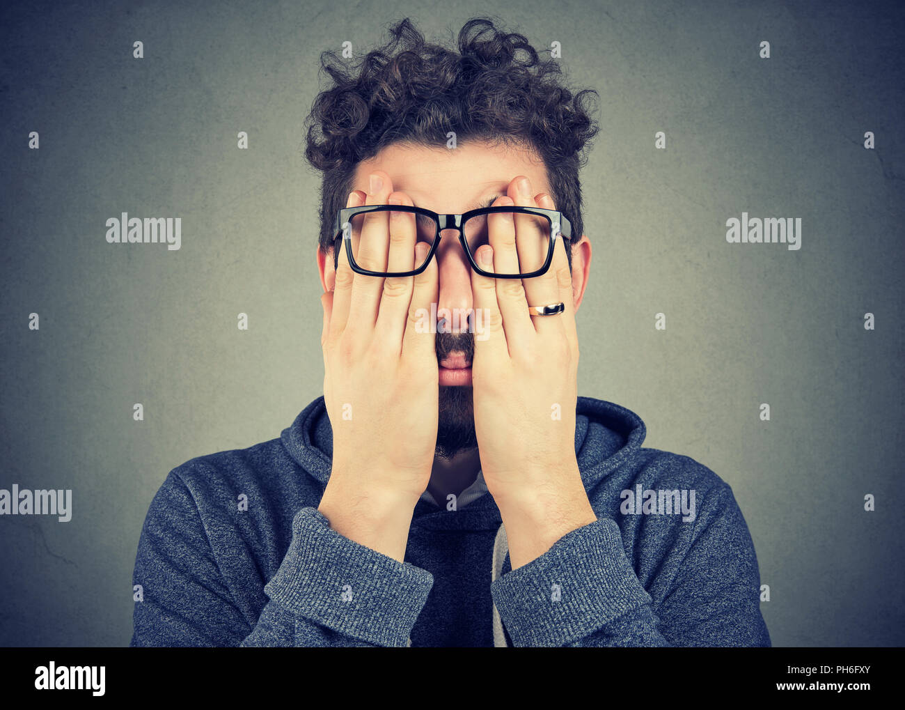 Closeup portrait young man in glasses covering face eyes with both hands isolated on gray wall background Stock Photo