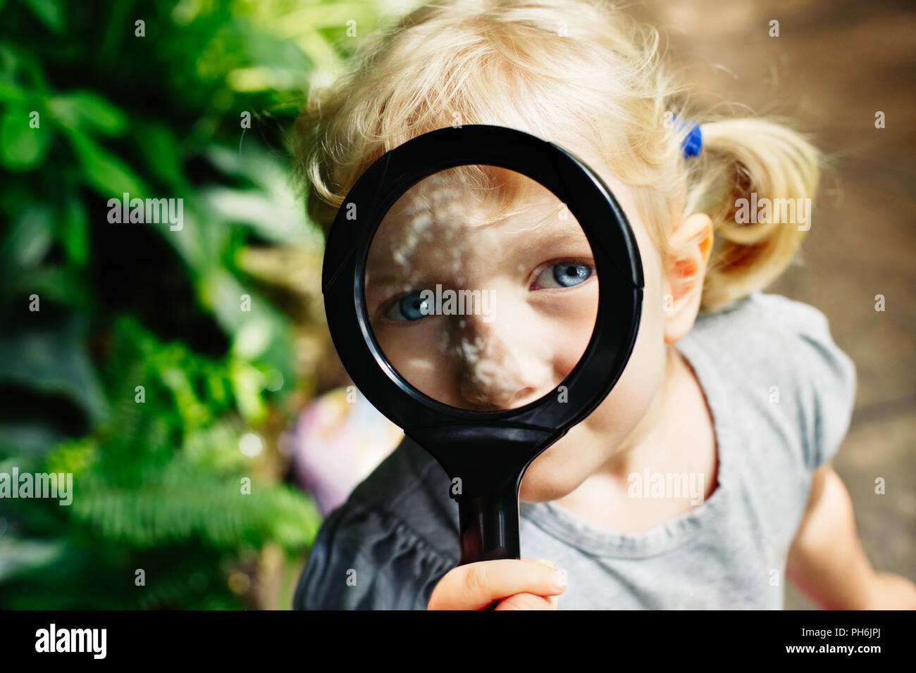 Portrait of cute adorable white Caucasian girl looking in camera through flowers through magnifying glass. Child with loupe studying learning nature.  Stock Photo