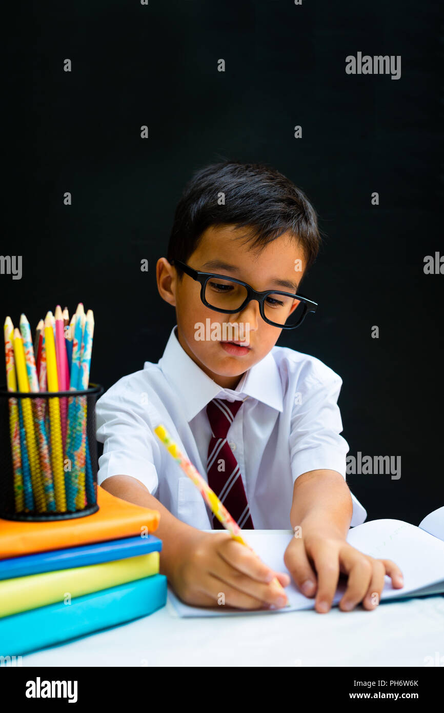 Cute smart handsome school boy in white shirt with eye glasses, writing at the desk in classroom, happy first grader pupil, smiling, knowledge study b Stock Photo