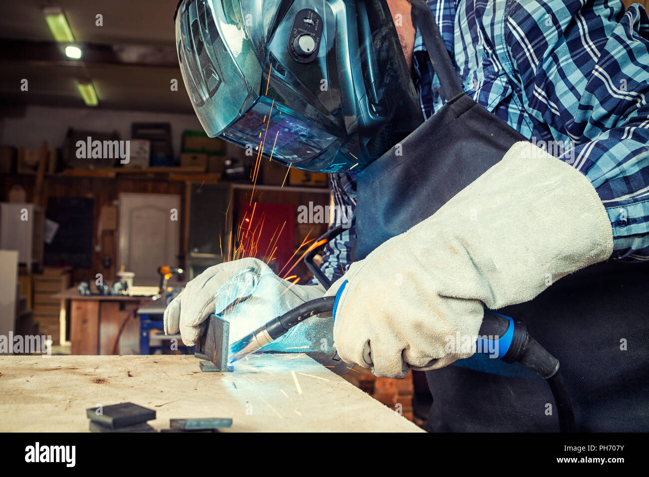 Close up of a young  man welder in  uniform, welding mask and welders leathers, weld  metal  with a arc welding machine  in workshop, blue and orange  Stock Photo