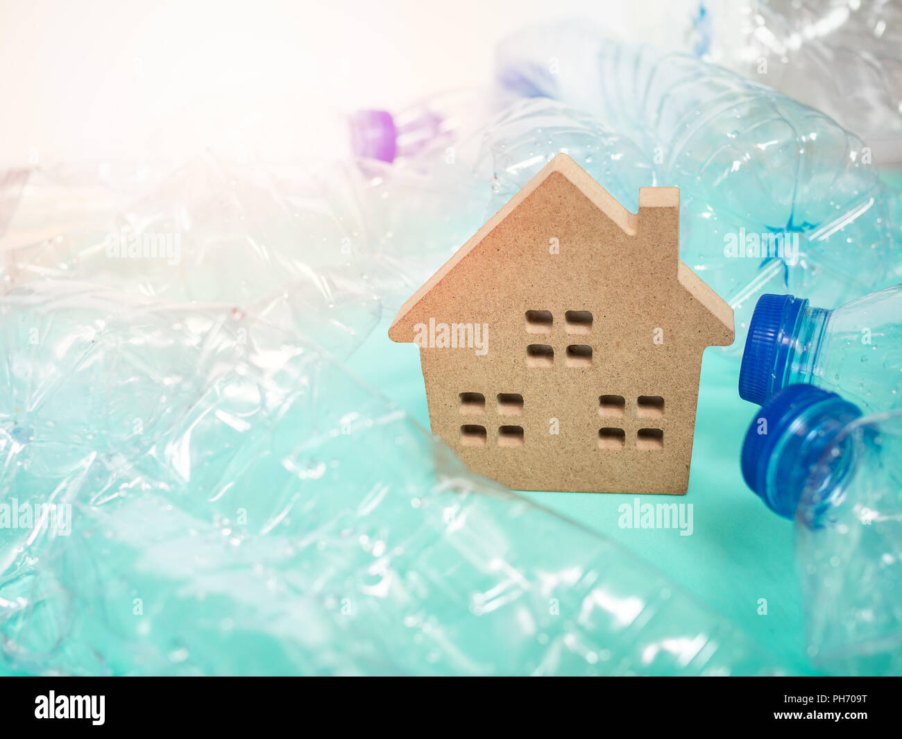 Wooden house and empty crushed recycle clear plastic drink water bottles on blue background with light and copy space. Global warming concept. Stock Photo