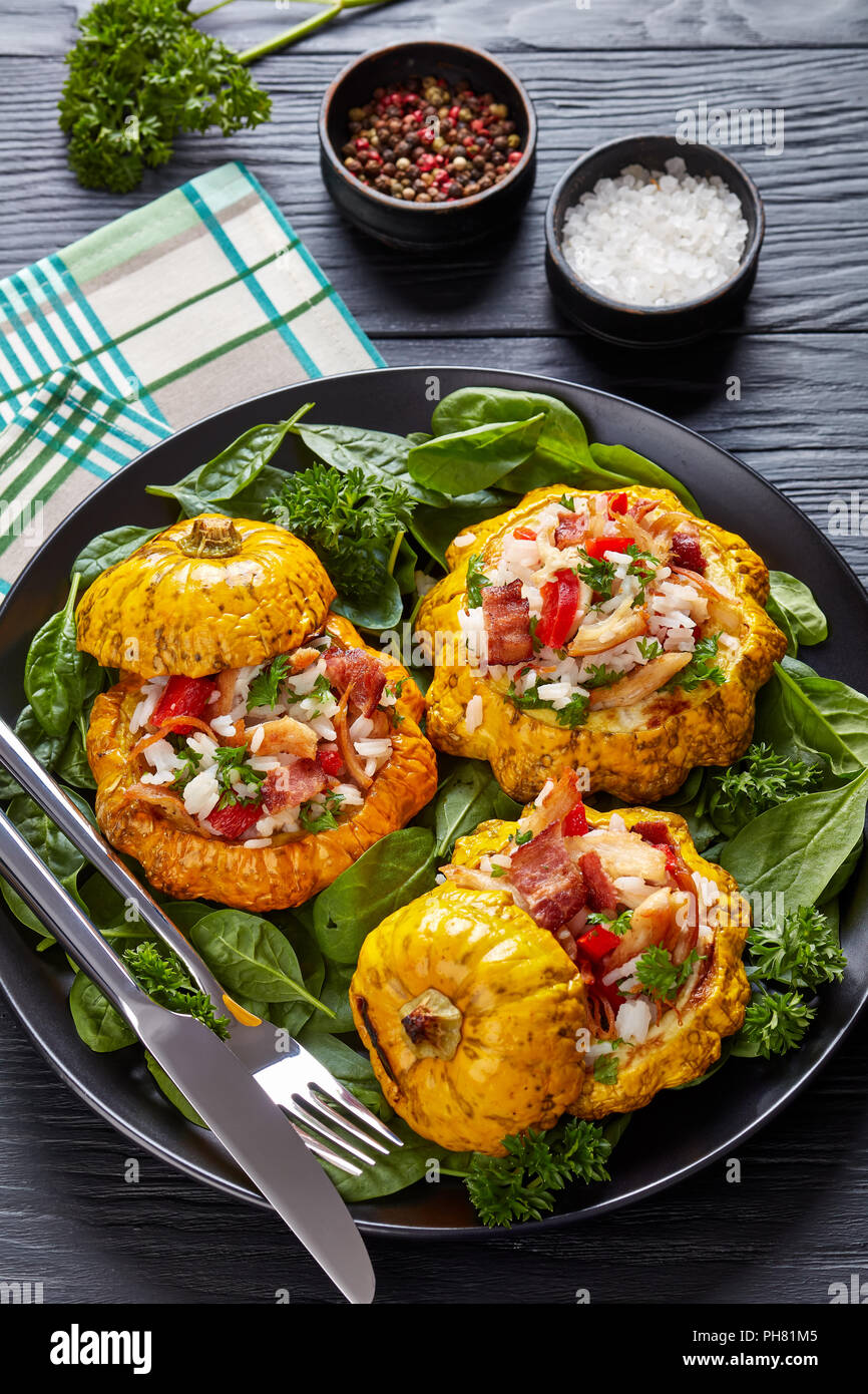 baked Pattypan squash stuffed with rice, fried chicken meat, crispy fried bacon, red bell pepper and served with spinach leaves and parsley on a black Stock Photo