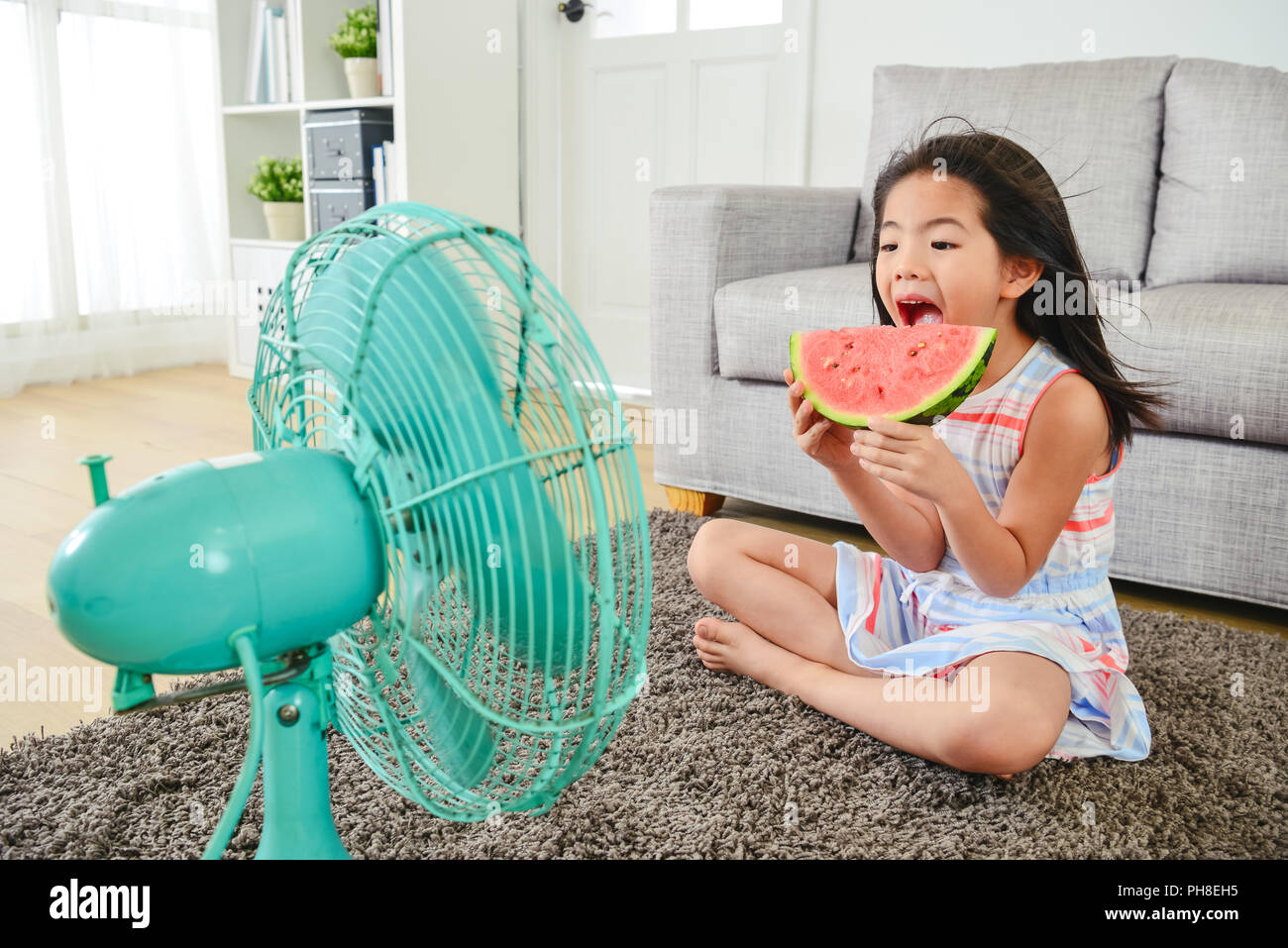 lovely girl eating melon with a big bite. sitting in front of a fan with legs crossed. cooling herself down from heat. Stock Photo
