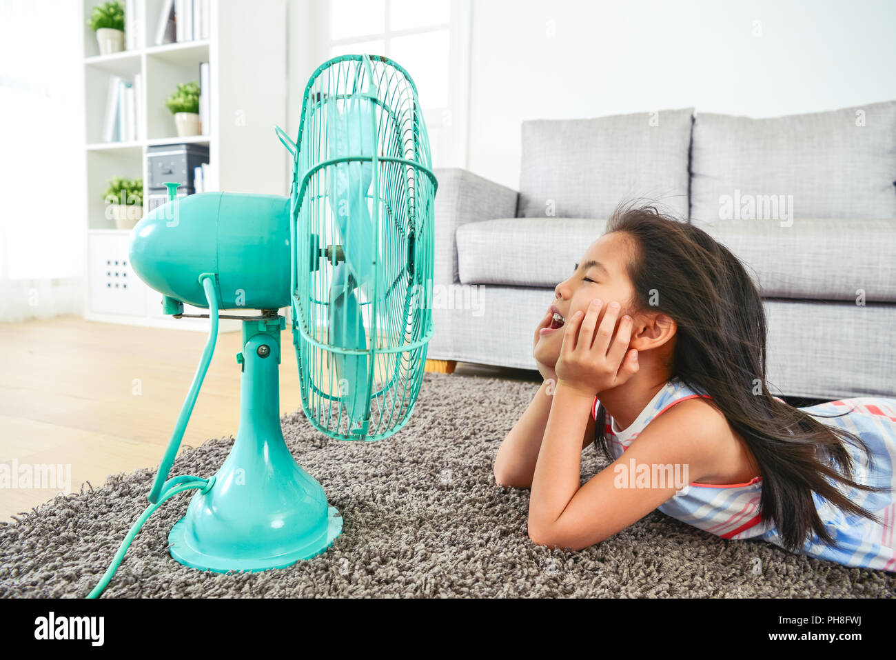 happy kid lying down in front of electric fan. with eyes clossed and hair blown by the breeze.  half-body view from side. Stock Photo