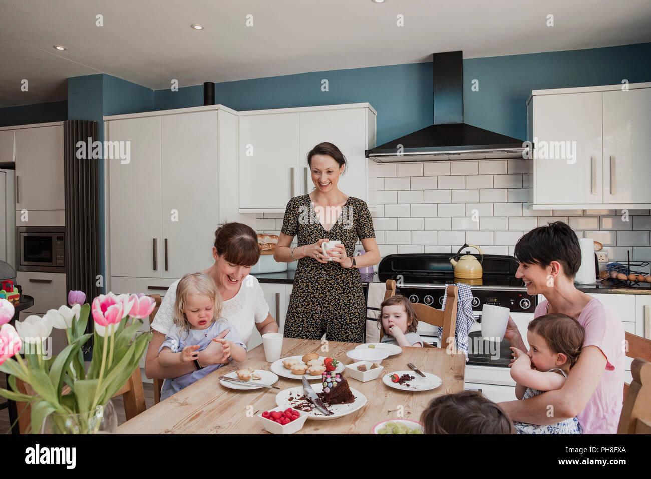 Three mid adult women in a kitchen with their daughters. Two of the women are sitting down with their daughters sitting on their laps while they eat t Stock Photo