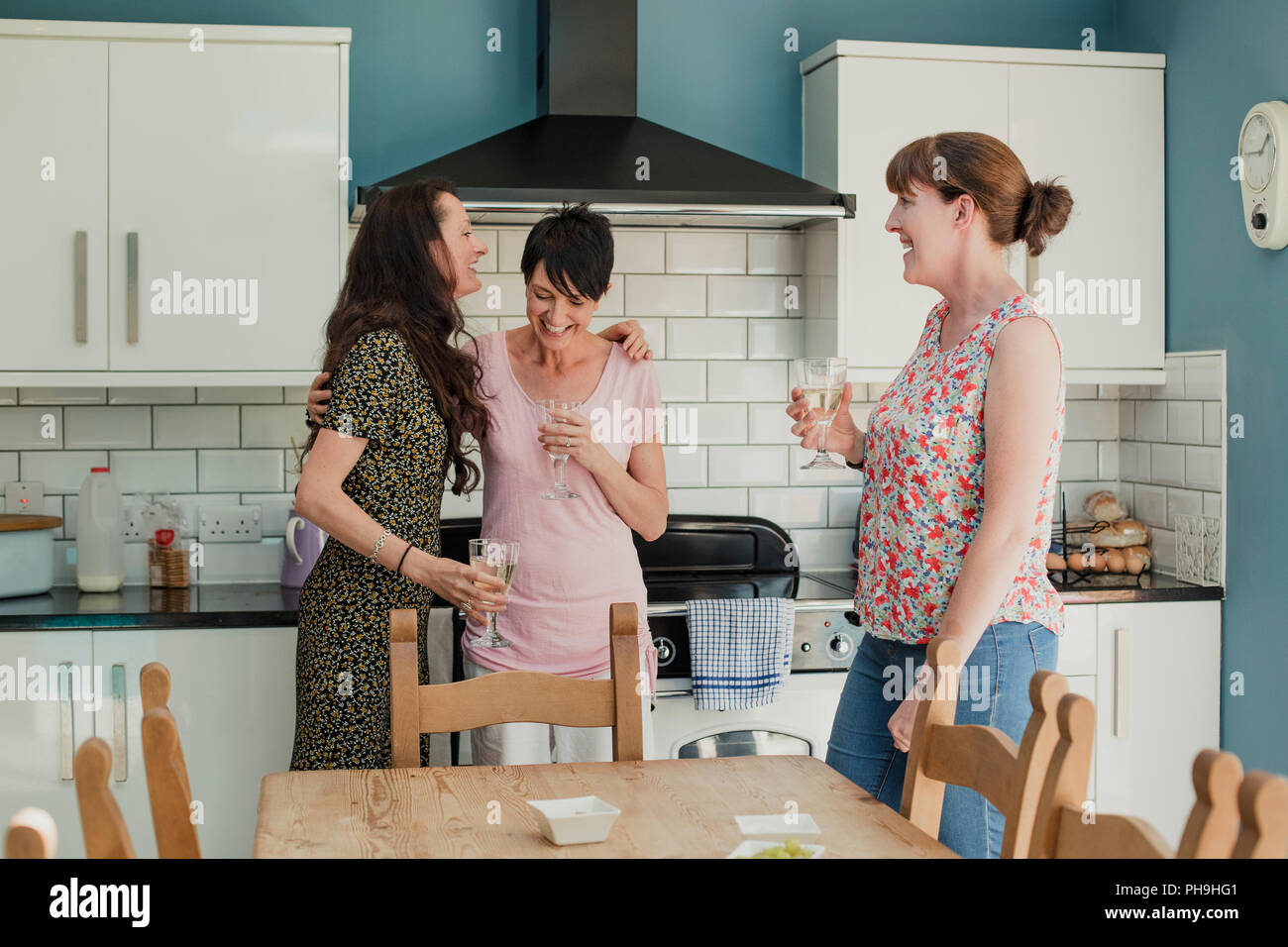 Three mid adult women standing in a kicthen enjoying a glass of wine. The women are all standing around the stove in the kitchen, talking and laughing Stock Photo