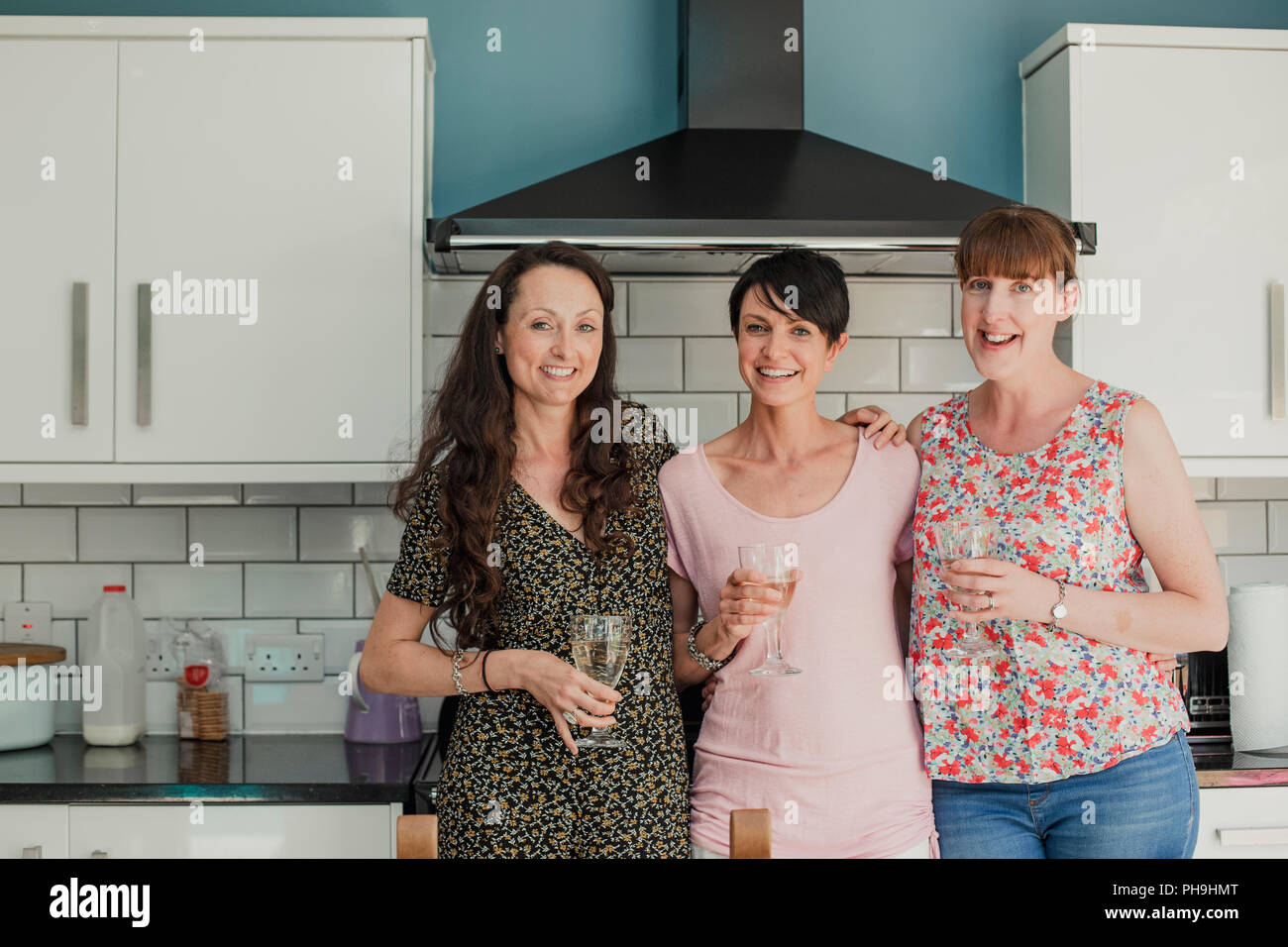 Three mid adult women standing in a kicthen enjoying a glass of wine. The women are all standing around the stove in the kitchen, looking at the camer Stock Photo