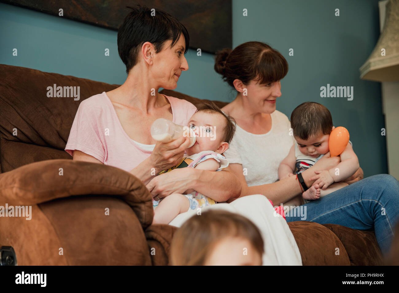 Mid adult mother is feeding her baby daughter a bottle of milk. sitting next to her is her mid adult friend with her baby boy sitting on her lap and w Stock Photo