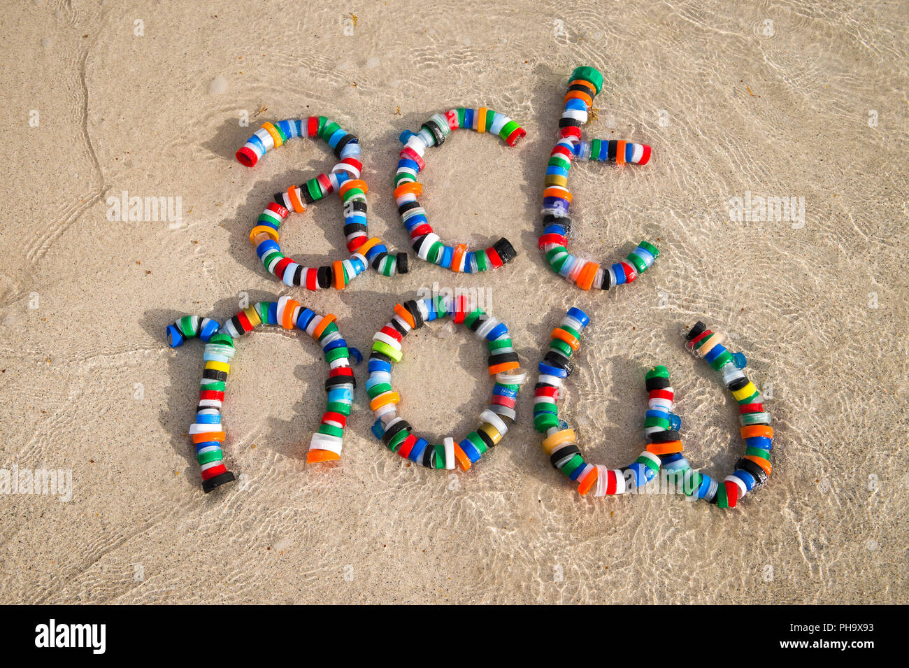 Colorful plastic bottle caps spell out 'act now' on a sandy beach as a wave laps around. Take action on pollution - reduce, reuse and recycle Stock Photo
