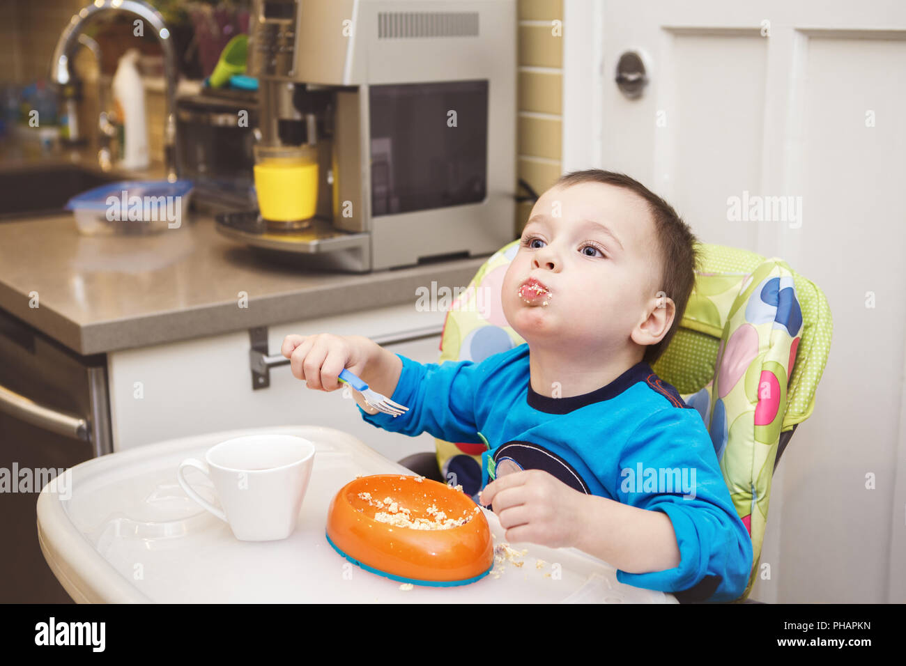 Portrait of cute adorable little toddler boy eating cottage cheese using fork, drinking orange juice, making funny face, sitting in high chair in kitc Stock Photo