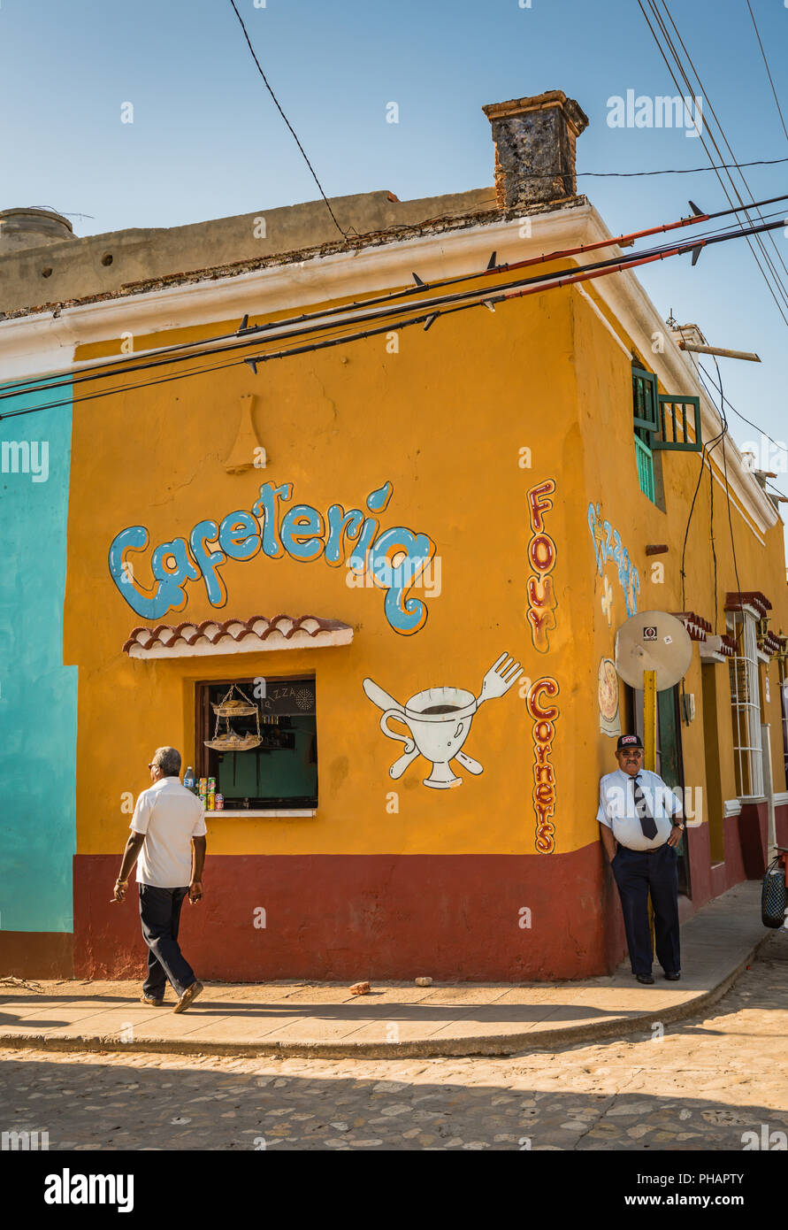 Trinidad, Cuba / March 15, 2016: Cuban man leans against colorful corner coffee shop with painted mural in UNESCO heritage city. Stock Photo