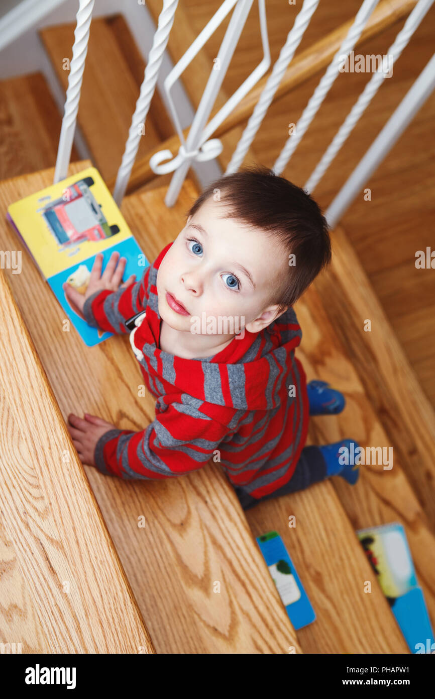 Portrait od cute adorable little boy toddler two years old with children books climbing on stair indoors inside looking up, shot overhead top above Stock Photo