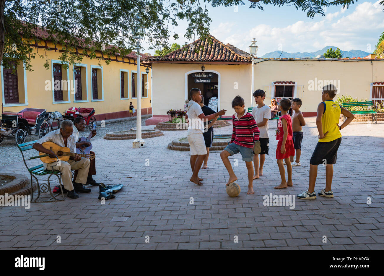 Trinidad, Cuba / March 15, 2016: Young boys playing soccer in a town square as two men play traditional Cuban music on guitars. Stock Photo