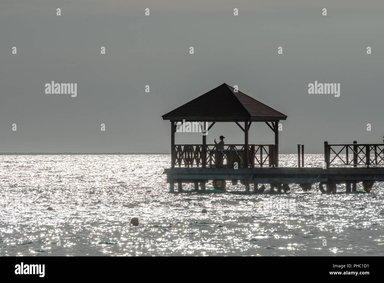 Bayahibe, Dominican Republic, 27 August 2018. A tourist uses her phone at a pier over the Caribbean sea. The grey look is due to the extreme backlight Stock Photo