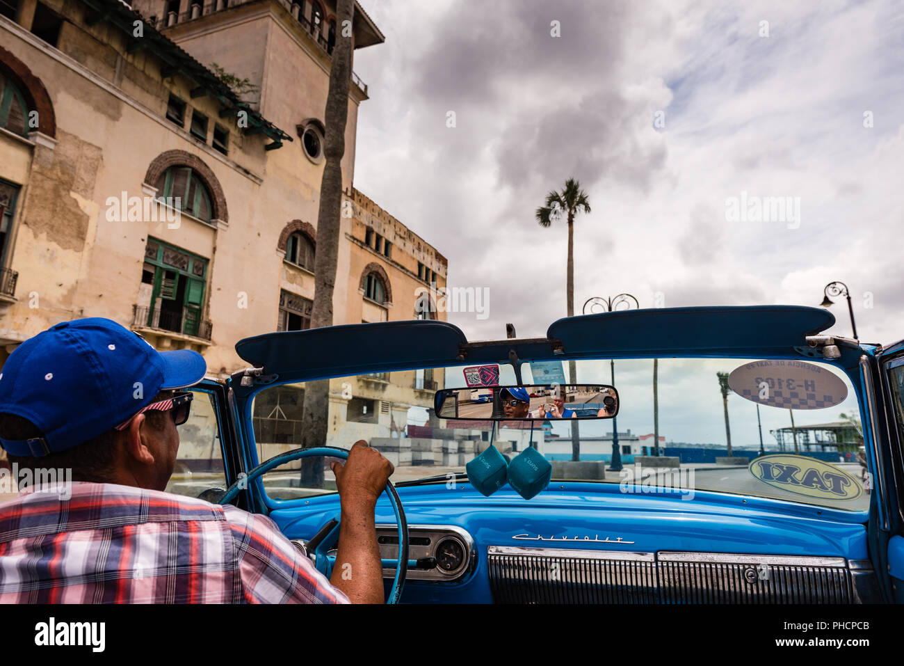 Havana, Cuba / March 22, 2016: Cuban man drives blue vintage convertible taxi under gray skies  through streets lined with Colonial architecture. Stock Photo