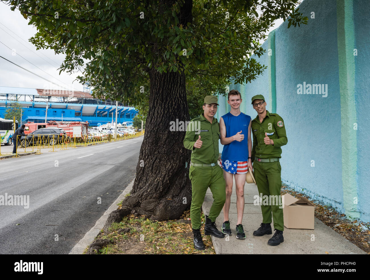 Havana, Cuba / March 22, 2016: American tourist, dressed in patriotic shorts, gives a thumbs up with two Cuban soldiers dressed in uniform fatigues on Stock Photo
