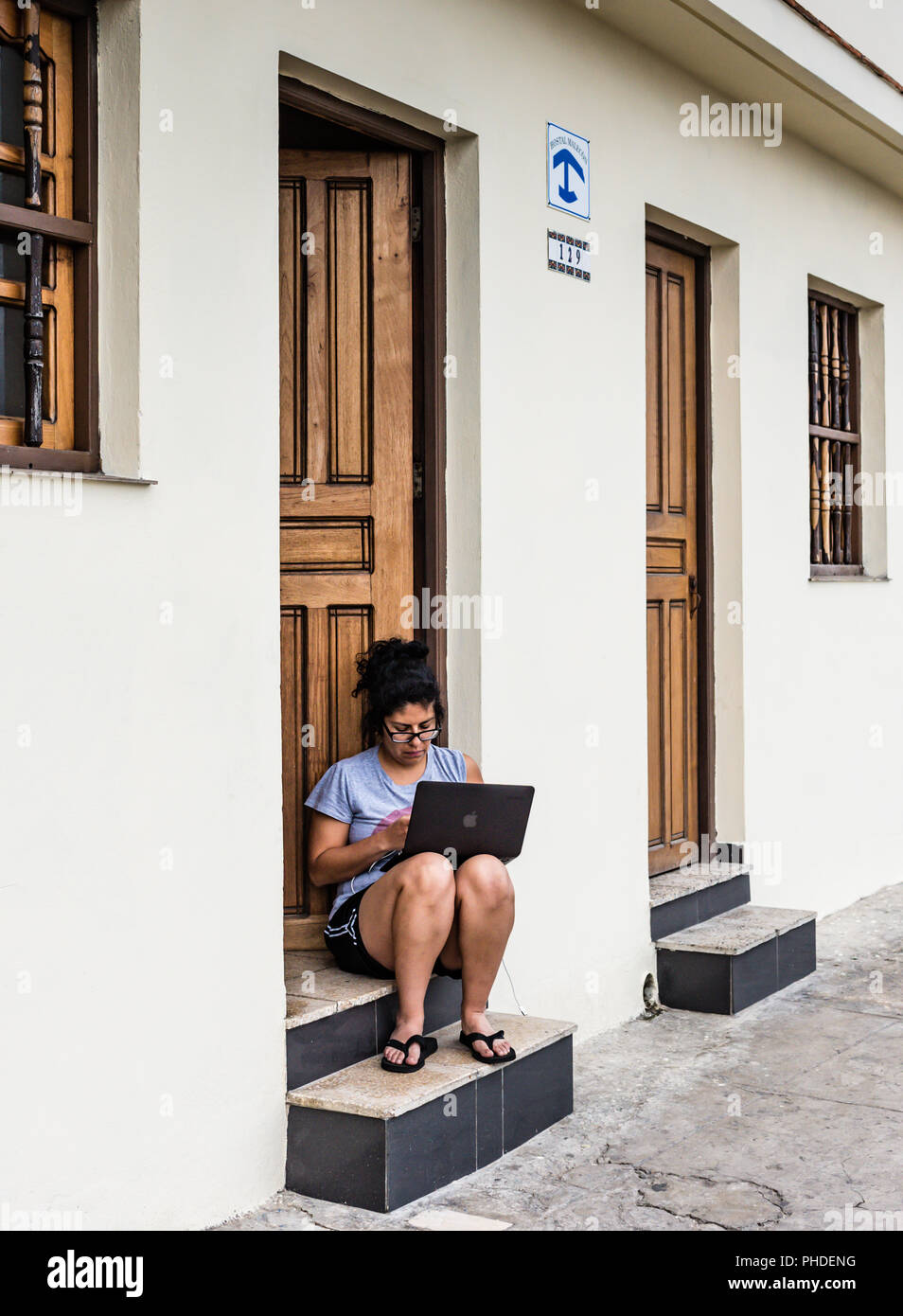 Havana, Cuba / March 21, 2016: Although Cuba has limited wifi connectivity, a woman works on a laptop at a hostel doorstep. Stock Photo