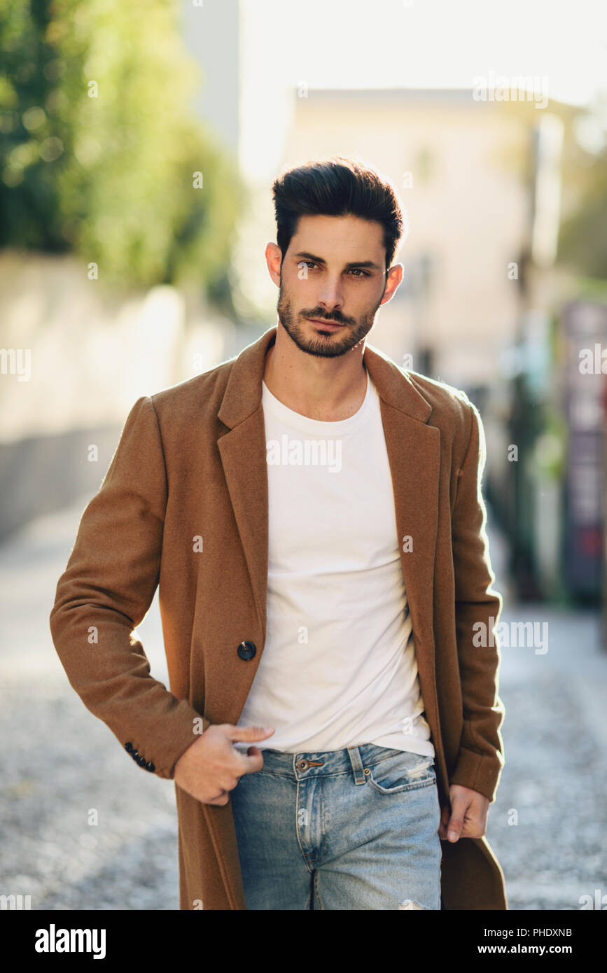 Young man wearing winter clothes in the street. Young bearded guy with  modern hairstyle with coat, blue jeans and white t-shirt Stock Photo - Alamy