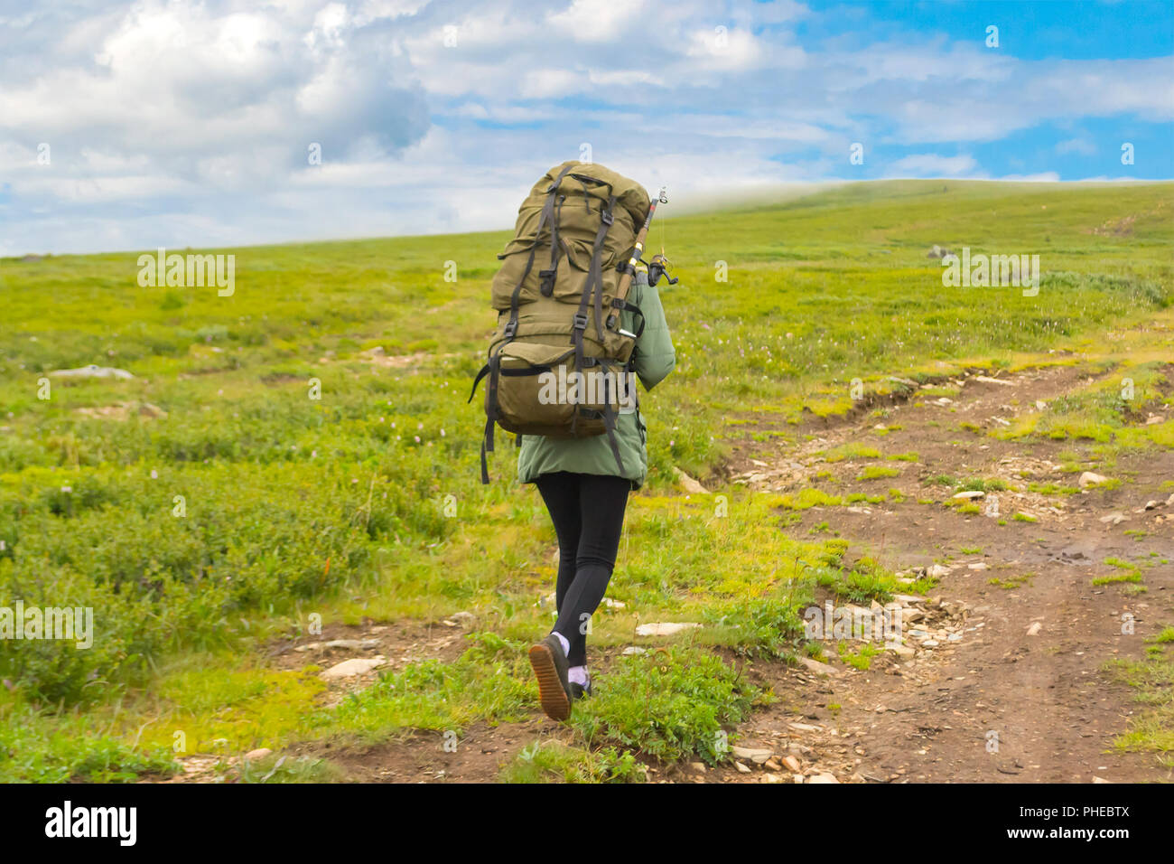 Young girl travel with big green backpack and fishing rod in it go on the road, along the green hill with green grass to the mountains and adventures  Stock Photo