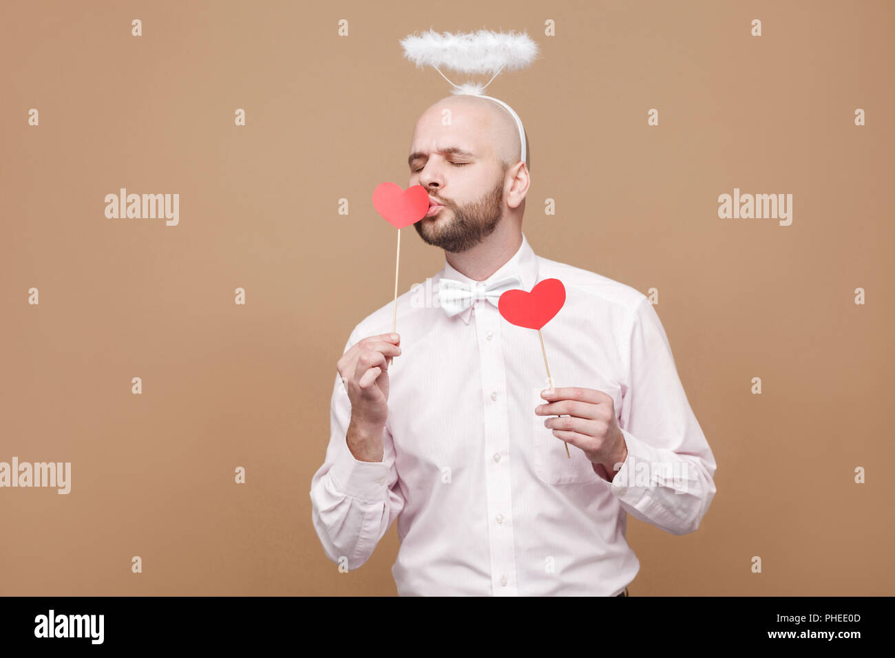 Happy handsome middle aged bald bearded angel in shirt and white halo on head standing and holding two red heart stickers and kissing one of them. stu Stock Photo