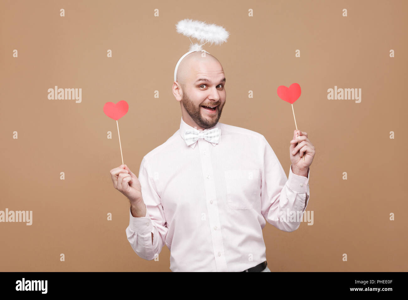 Happy handsome middle aged bald bearded angel in shirt and white halo on head standing and holding two red heart stickers and smiling. indoor studio s Stock Photo